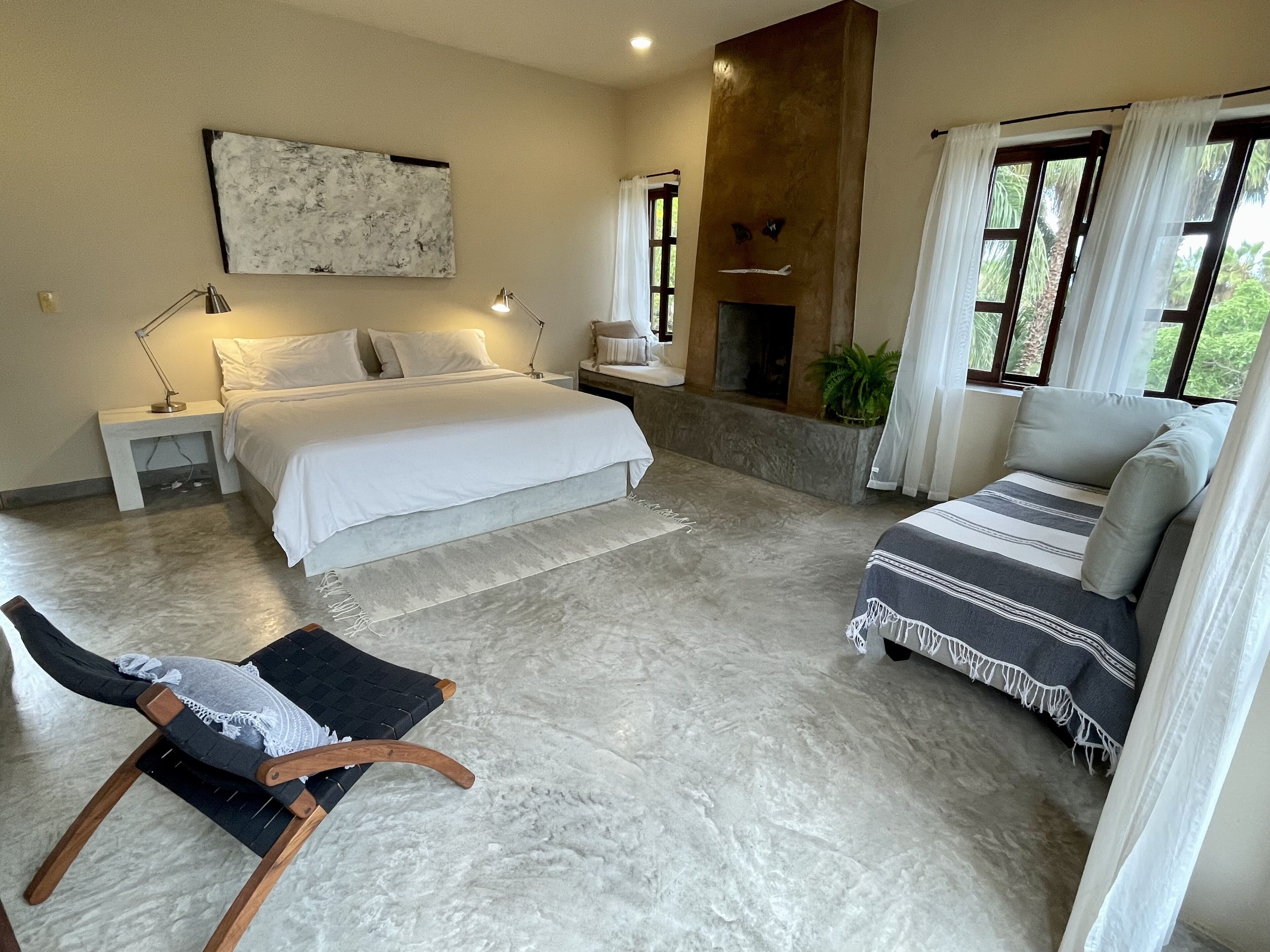 Une chambre à coucher comprenant un lit, des tables de chevet, une chaise, un canapé, une cheminée et une vue sur la verdure par les fenêtres.