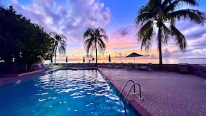 A pool area with lounge chairs and umbrellas, surrounded by palm trees, with a view of the sea and a sunset sky. - Stunning Ocean View Condo steps to 7 Mile Beach-Ground Floor! (George Town)