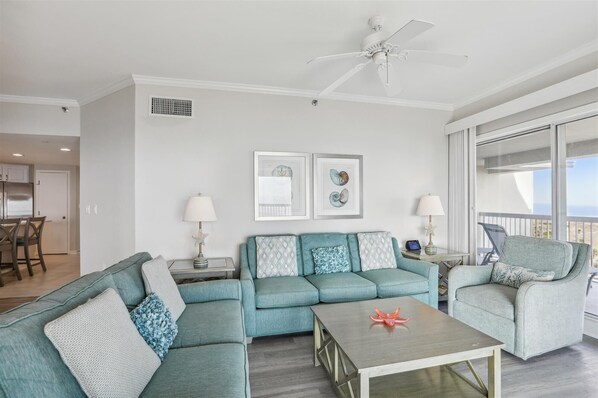 A living room with a sectional sofa, a coffee table, and a ceiling fan. There are two framed pictures on the wall, a window with a view, and a balcony with chairs.