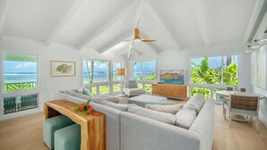 Ett rymligt vardagsrum med ett stort fönster med utsikt över havet, en takfläkt och ett träbord som används som soffbord, dekorerat med en blomsteruppsättning.