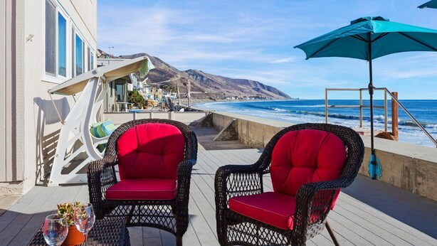 Ein Balkon mit Korbstühlen und einem Tisch, mit Blick auf die Küste und Berge im Hintergrund.