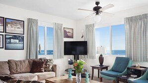 Ein Wohnzimmer mit einer Couch, Sesseln, einem Couchtisch und einem Fernseher. Vorhanden sind Vorhänge, ein Deckenventilator und Meerblick durch große Fenster.
