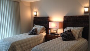A hotel room with two beds, each with a headboard, a bedside table with a lamp, and a window with blinds.