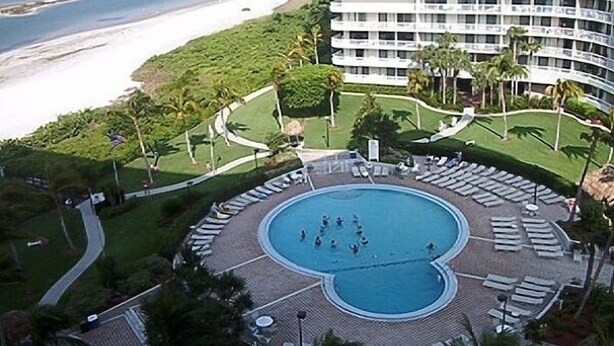 A hotel with a circular pool surrounded by lounge chairs, a beach view, and a multi-story building in the background.