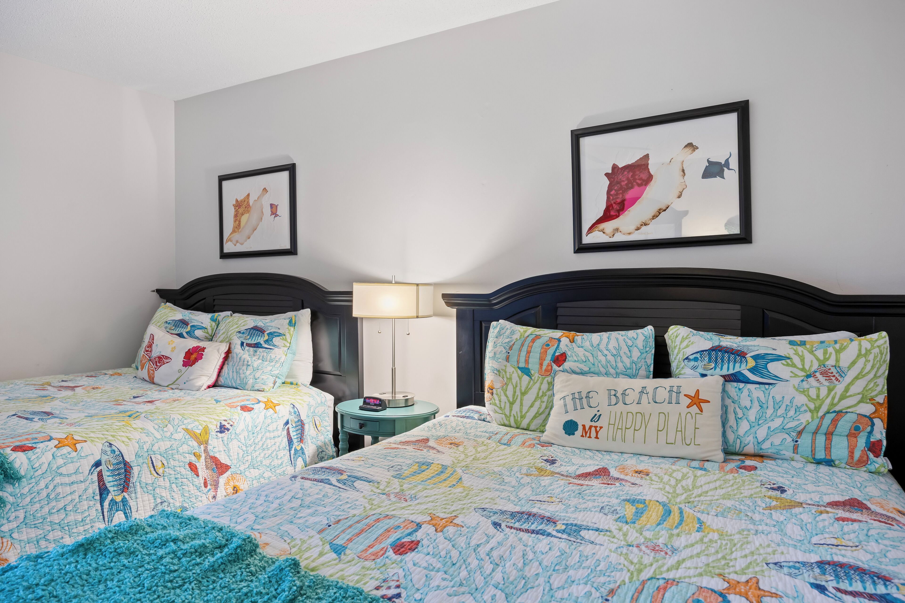 A bedroom with two beds, each with a colorful fish-themed bedspread, a nightstand with a lamp, and framed artwork on the walls.