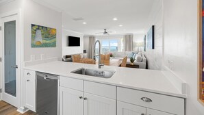 A modern kitchen with white cabinetry, a built-in dishwasher, and a large island with a sink. There is a painting on the wall and a ceiling fan in the dining area. - Gulf-front 30A Townhome -  Steps to the Beach.
FALL IS RIGHT AROUND THE CORNER.. (Seagrove Beach)
