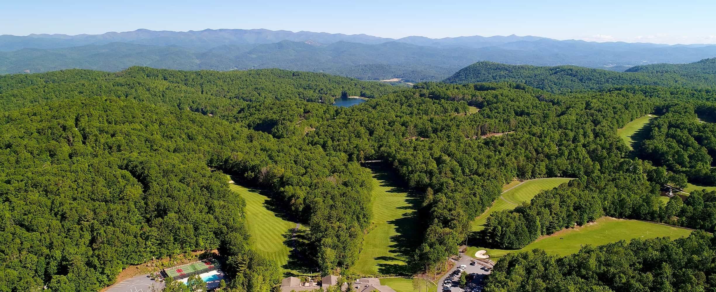 An aerial view of a forested area with a golf course, a lake, and distant mountains.