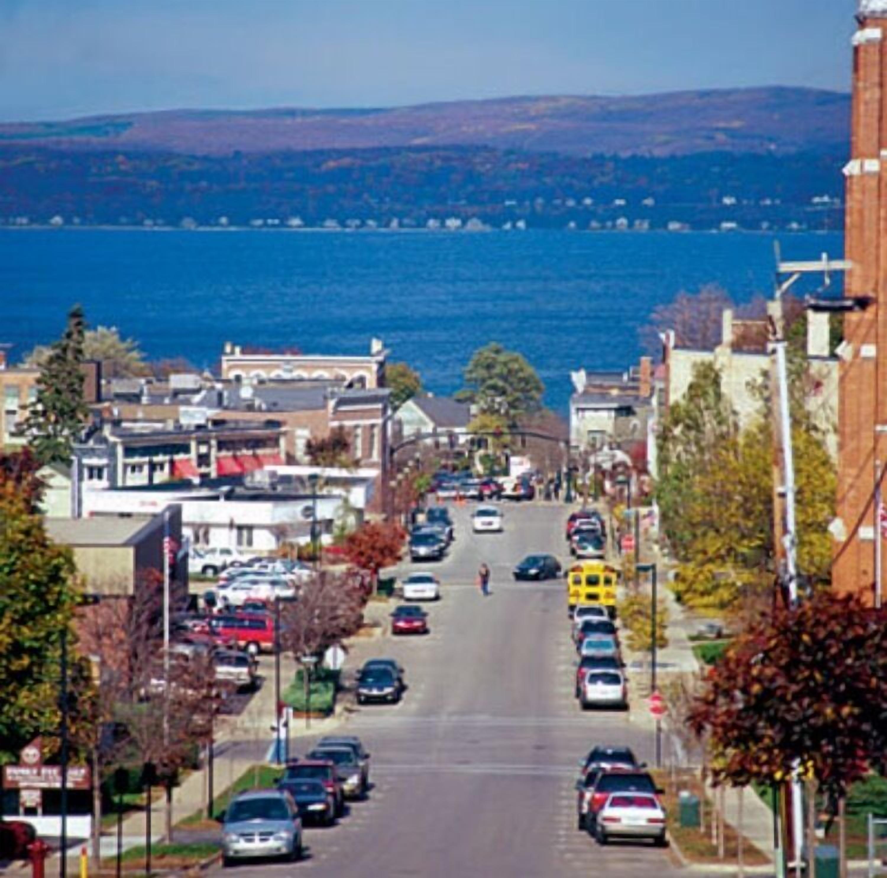 A street view with buildings, parked cars, and a body of water in the background.