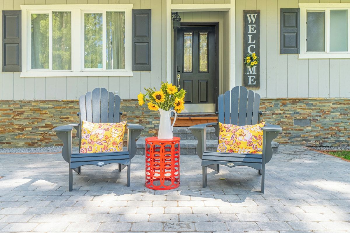 Zwei Adirondack-Stühle mit blumigen Kissen vor einem Haus, an dem ein 'WELCOME'-Schild hängt.