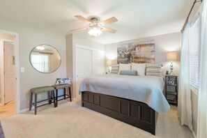 A bedroom with a bed, a ceiling fan, a round mirror, a bench, and a nightstand with a lamp.