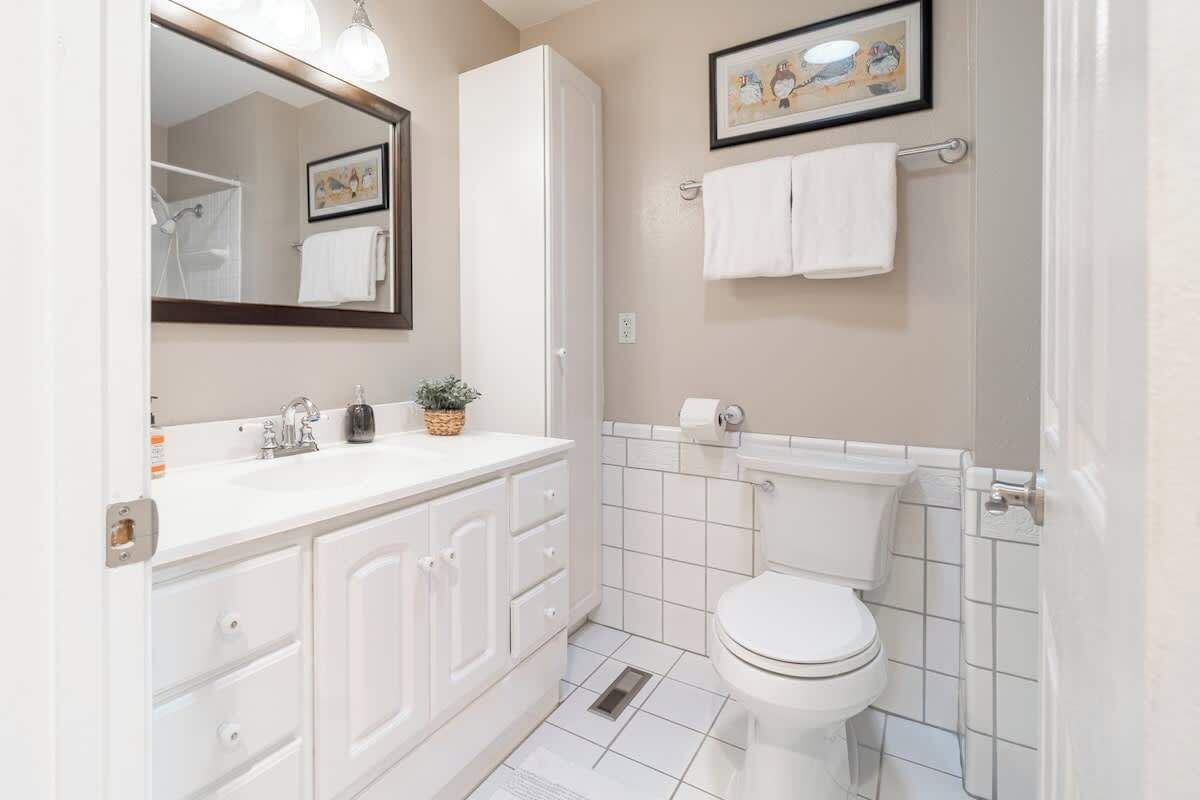 A modern bathroom with a white sink, vanity, and toilet. There is a mirror with lights, a framed picture on the wall, and a small plant on the vanity.