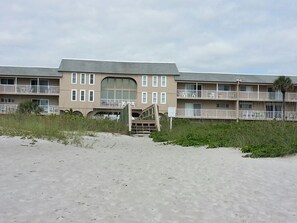 Un hotel frente al mar con entrada central, varios balcones y una playa de arena en primer plano.