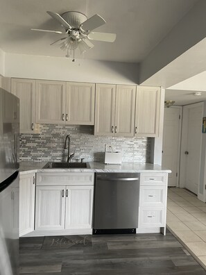 Una cocina con armarios de madera clara, salpicadero con azulejos grises, lavavajillas de acero inoxidable y ventilador de techo.