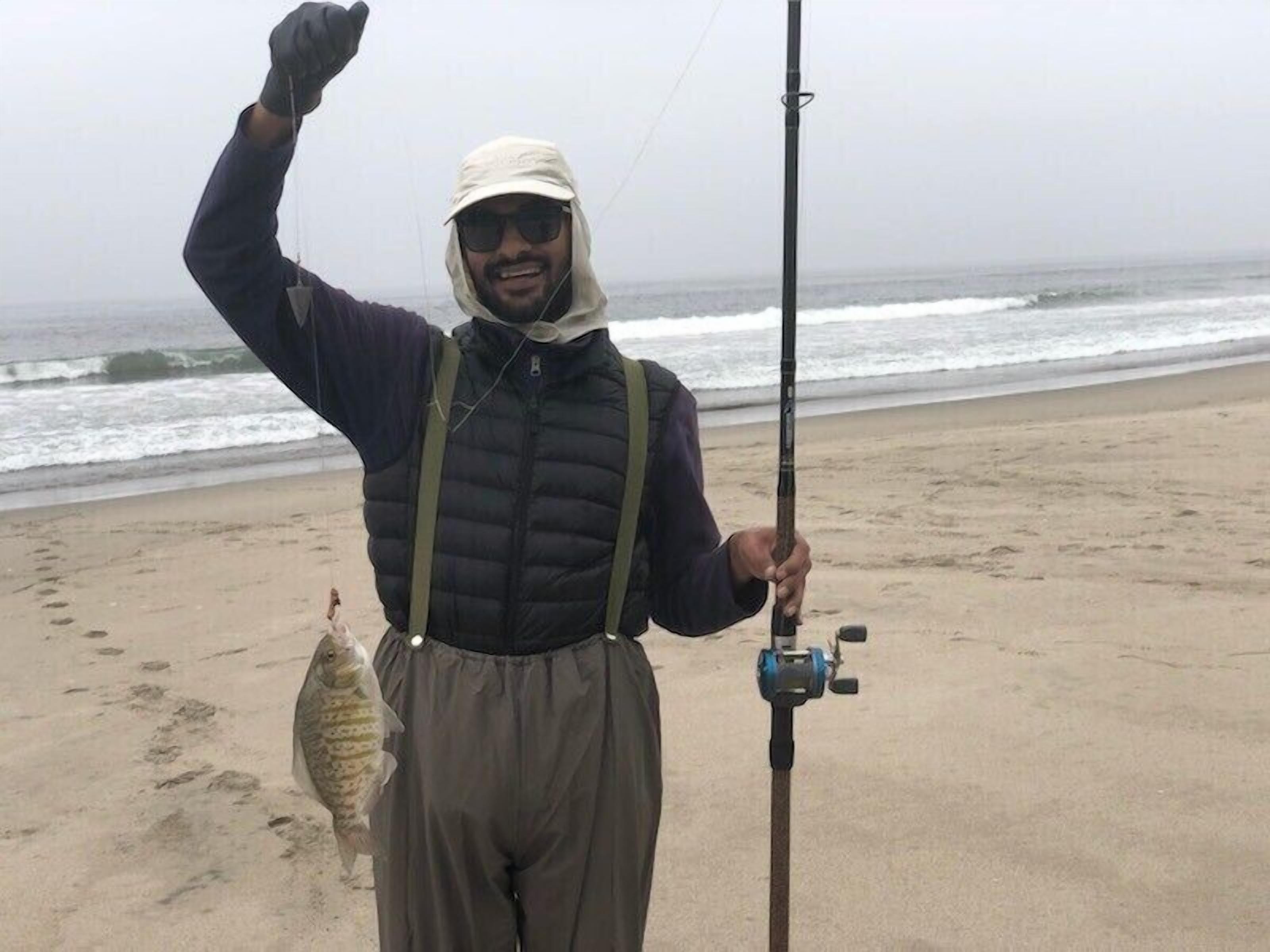 Eine Person steht am Strand, hält eine Angel und einen Fisch, im Hintergrund das Meer.