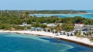 A coastal area with a beach, clear blue waters, and a line of wind turbines in the background. - Enjoy Caribbean weather in the USA. Openings August 2026 forward!  (Cudjoe Key)