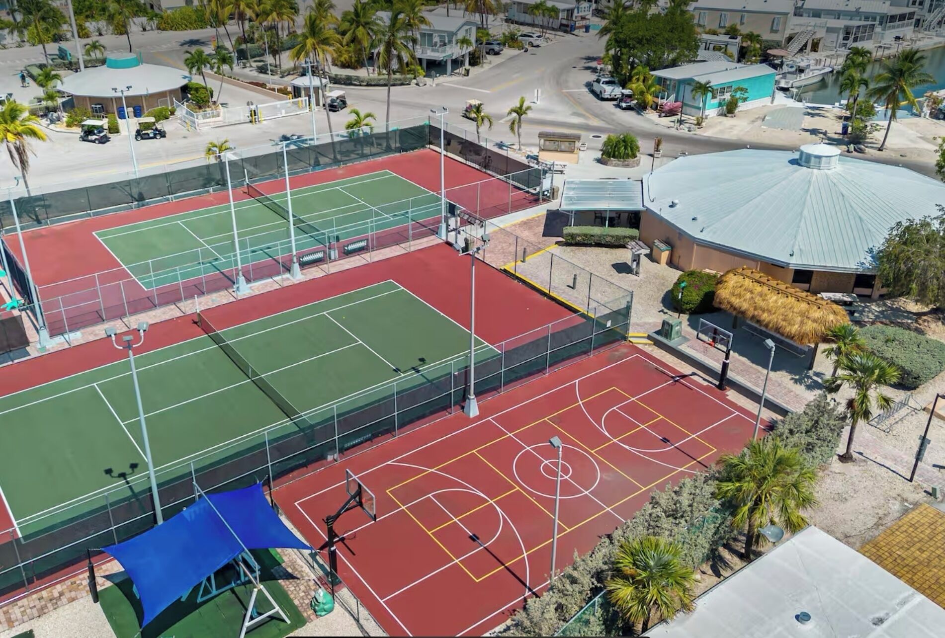 An aerial view of a sports complex with multiple tennis courts, a covered seating area, and surrounding buildings.