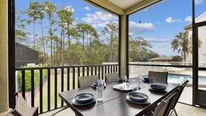 Un espacio de comedor al aire libre con techo, con una mesa dispuesta para cuatro, con vista a una piscina y paisajes de árboles y casas.