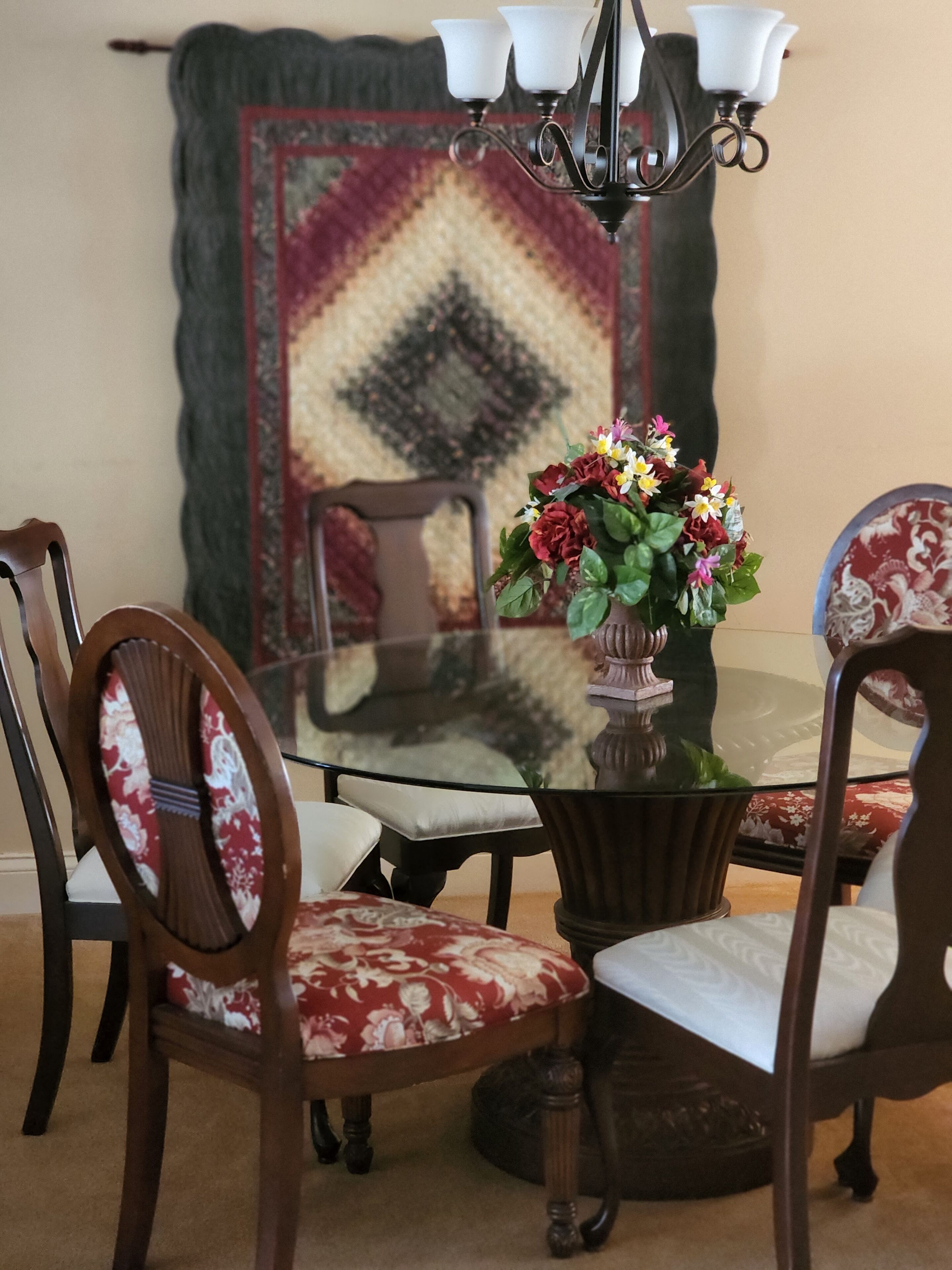 Uma mesa de jantar com tampo de vidro, rodeada por cadeiras de madeira com estofados florais. Um vaso com flores está sobre a mesa.