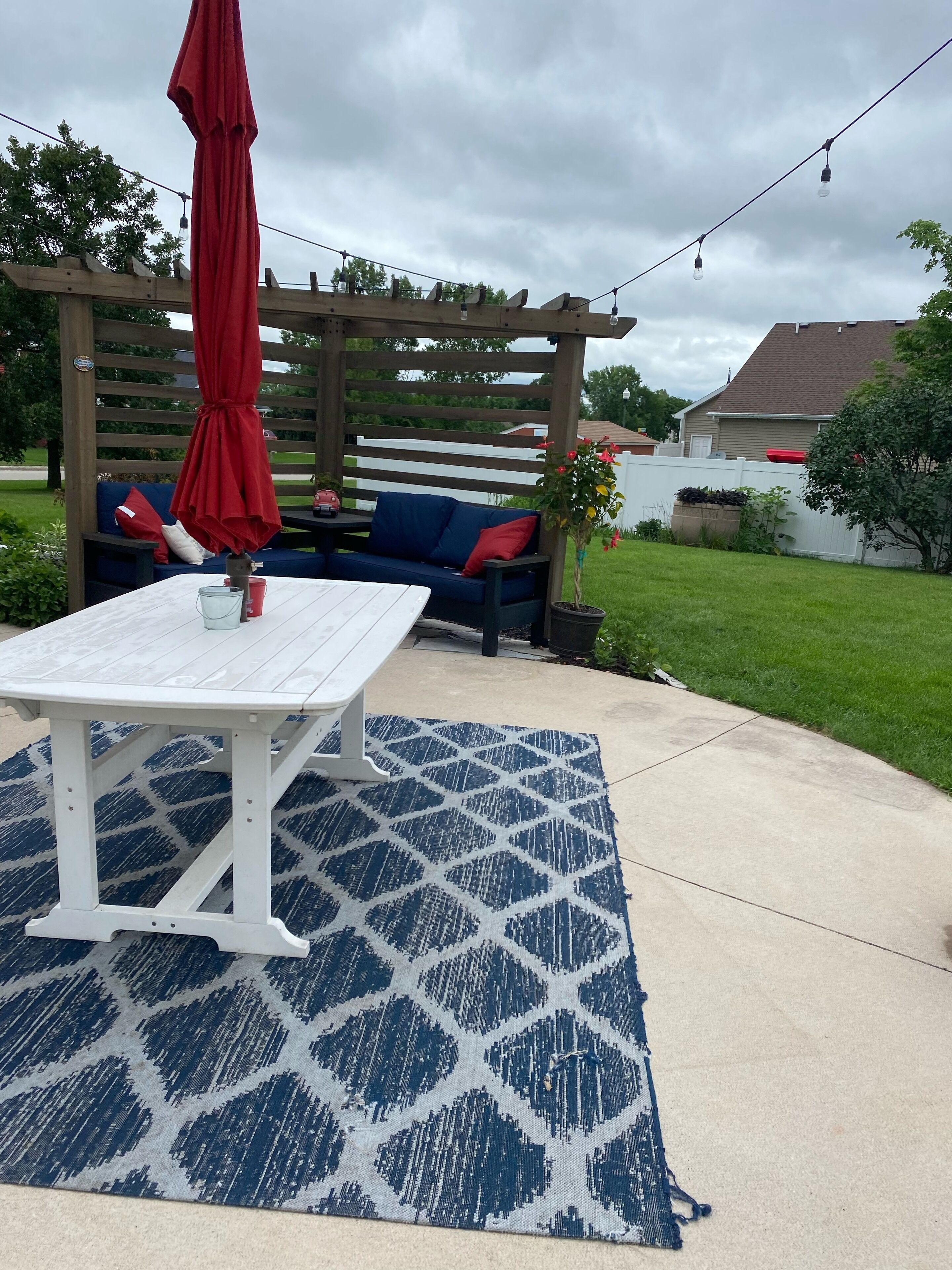 Une terrasse extérieure avec une table blanche et un tapis à motifs bleus, une pergola en bois et un canapé bleu agrémenté de coussins rouges.