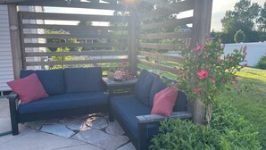 A covered patio with a blue sofa and red cushions, a small table with a red toy car, and a potted plant with pink flowers. - Friendly, Quiet Cul-de-sac - 18 Minutes from Oshkosh EAA Air Show Appleton ATW! (Neenah)