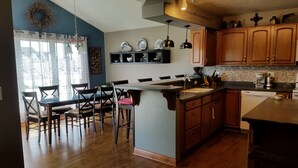 A kitchen with wooden cabinets, a dining area with chairs, a built-in island with a sink, and a decorative wreath on the wall. - Friendly, Quiet Cul-de-sac - 18 Minutes from Oshkosh EAA Air Show Appleton ATW! (Neenah)