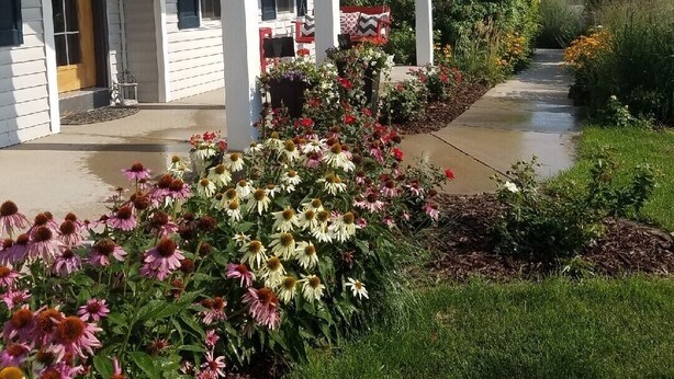 A white building with a porch, American flags, and a garden with flowers. - Friendly, Quiet Cul-de-sac - 18 Minutes from Oshkosh EAA Air Show Appleton ATW! (Neenah)