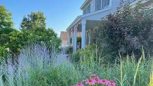 A residential house with a porch, surrounded by lush greenery and flowering plants. - Friendly, Quiet Cul-de-sac - 18 Minutes from Oshkosh EAA Air Show Appleton ATW! (Neenah)