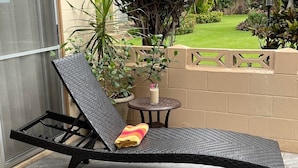 Una silla de lounge negra con cojín y una toalla, ubicada en un patio con baldosas, junto a una pequeña mesa redonda y una planta en maceta.