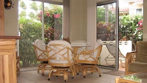 Un comedor con sillas de mimbre y una mesa de vidrio, puertas correderas de cristal que dan a un jardín con plantas tropicales.