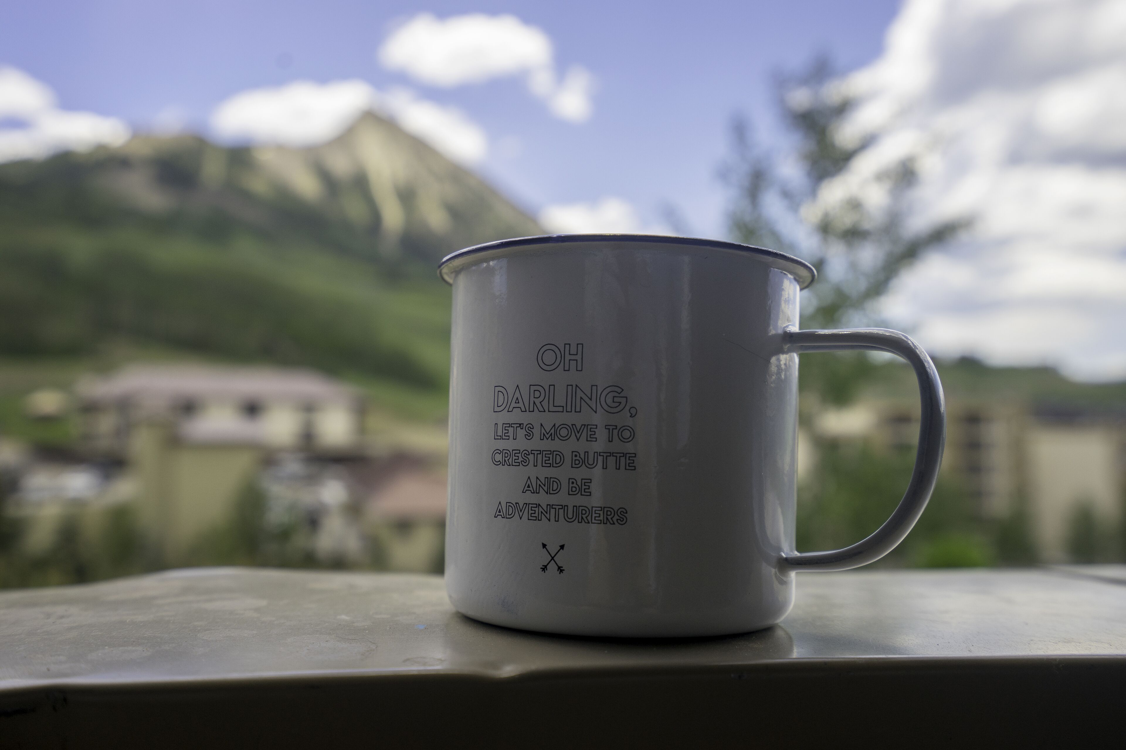 Una taza blanca con texto sobre una mesa, con una montaña y edificios al fondo.