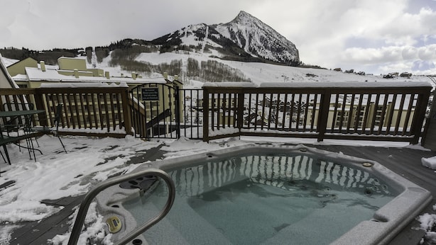 Ein Whirlpool auf einer Holzterrasse, umgeben von Schnee, mit einem Schild, das „Nachhaltigkeit! Bitte Whirlpool abdecken, wenn er nicht genutzt wird.“ steht. Im Hintergrund ist ein Berg zu sehen.