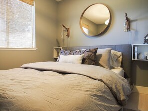 A neatly made bed with a large round mirror above it, a bedside table with a lamp, and a shelf with books and a decorative item.
