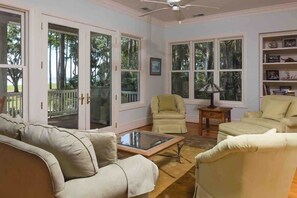 A spacious living room with a beige sofa, two armchairs, a glass coffee table, and a ceiling fan. Large windows provide a view of the outdoors.