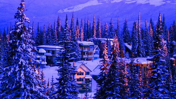 A snow-covered mountain resort with multiple buildings, including a lodge and cabins, surrounded by dense evergreen trees.