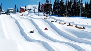 A snow-covered ski slope with multiple lanes for snow tubing, a ski lift in the background, and buildings at the top of the hill.