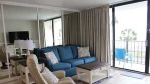 Un salon avec un canapé bleu, un fauteuil beige, une table basse, un téléviseur posé sur un meuble et un balcon avec une chaise bleue.