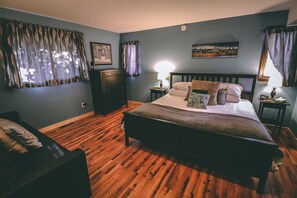 Une chambre avec un plancher en bois, un lit recouvert de plusieurs oreillers, une commode et une table de chevet avec une lampe.