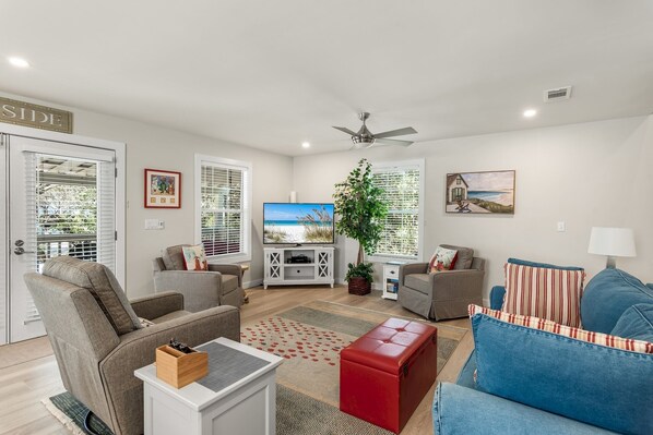 A living room with a TV, two sofas, a red ottoman, and a large plant. - Raised Cottage 'Dune Fine'- Easy Walk to Beach - Dogs Welcome! (Santa Rosa Beach)