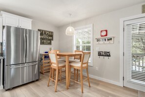 Ett kök med ett kylskåp i rostfritt stål, ett matbord och stolar i trä. Det finns dekorationer på väggarna, bland annat en skylt med texten ”FISK OCH OSTRON, DAGLIGEN FÄRSKT” och en skylt med texten ”LÅT OSS GÅ PÅ EN PROMENAD”.