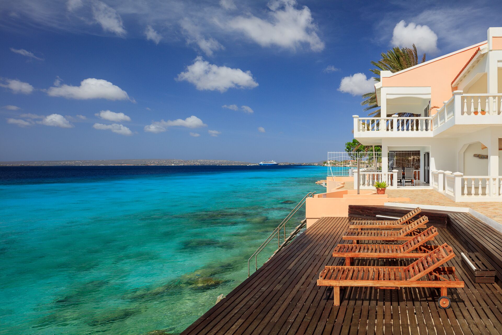 A wooden deck with lounge chairs overlooking turquoise waters, a white building with balconies, and a clear sky.