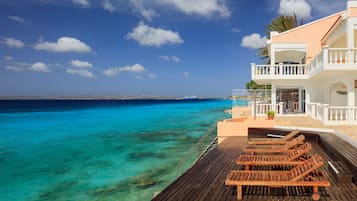 A wooden deck with lounge chairs overlooking turquoise waters, a white building with balconies, and a clear sky.