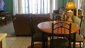 A living room with a brown sofa, a wooden dining table with chairs, a lamp, and a TV on a stand. - Check out new rates for summer (Panama City Beach)
