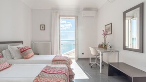 A hotel room with two beds, a desk, a bench, a window with a view of the sea, and an air conditioning unit.