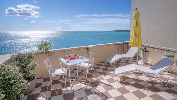 A terrace with a checkered floor, a table with chairs, a yellow umbrella, and lounge chairs overlooking a body of water.