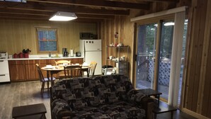 Acogedor interior de cabaña con sofá, mesa de comedor, sillas, zona de cocina y puerta corrediza de vidrio que da a una terraza.