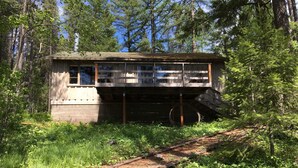 Una cabaña con terraza de madera y un espacio cubierto, rodeada de un bosque frondoso y un sendero de tierra.
