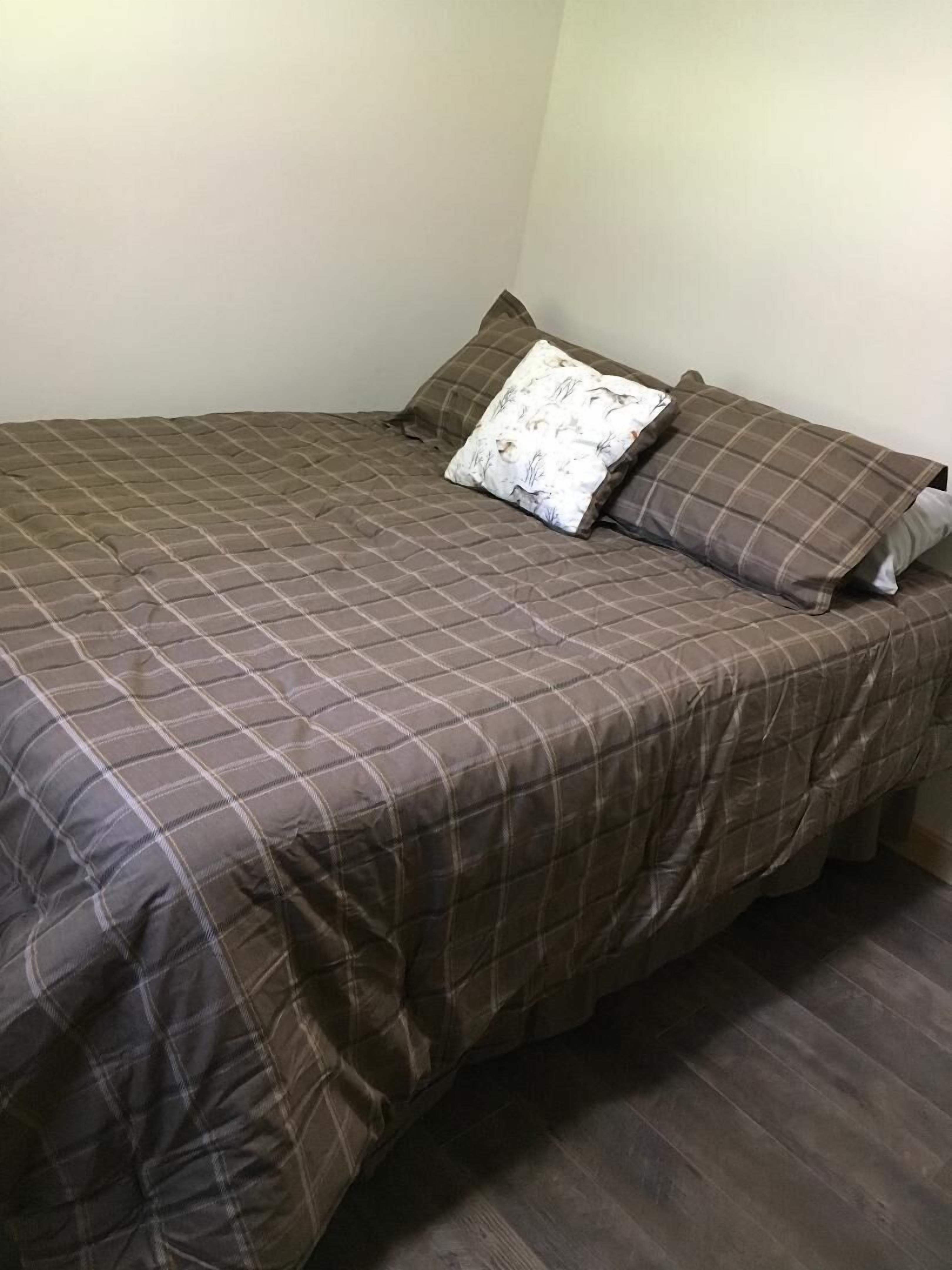 Une chambre avec un lit simple, une couverture à carreaux marron, deux oreillers et un mur blanc uni.
