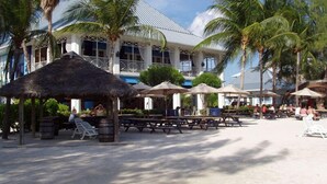 A beachside resort with thatched-roof structures, palm trees, and a large building with a white facade and blue accents. - The Pools #1- the Luxury Beachfront Hideaway with Private Pool (North Side, Cayman Kai,)