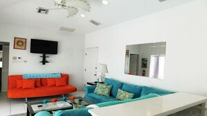 A modern living room with a white sofa, a teal sofa, and a white coffee table. There is a flat-screen TV mounted on the wall and a mirror above the teal sofa. - The Pools #1- the Luxury Beachfront Hideaway with Private Pool (North Side, Cayman Kai,)