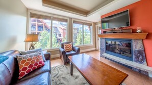 A living room with a fireplace, a television, and a sofa with patterned cushions. - Mountain Views - Canmore Luxury Condo in Solara Resort - Steps to restaurants (Canmore)